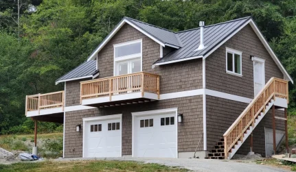 A newly constructed two-story brown house with a dark metal roof, two white garage doors, and two wooden balconies.
